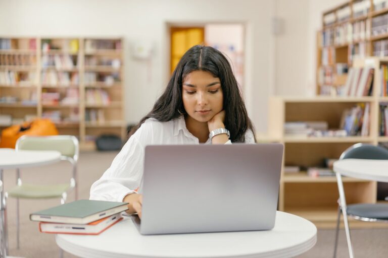 A young woman in a library studying online with a laptop and books around her.